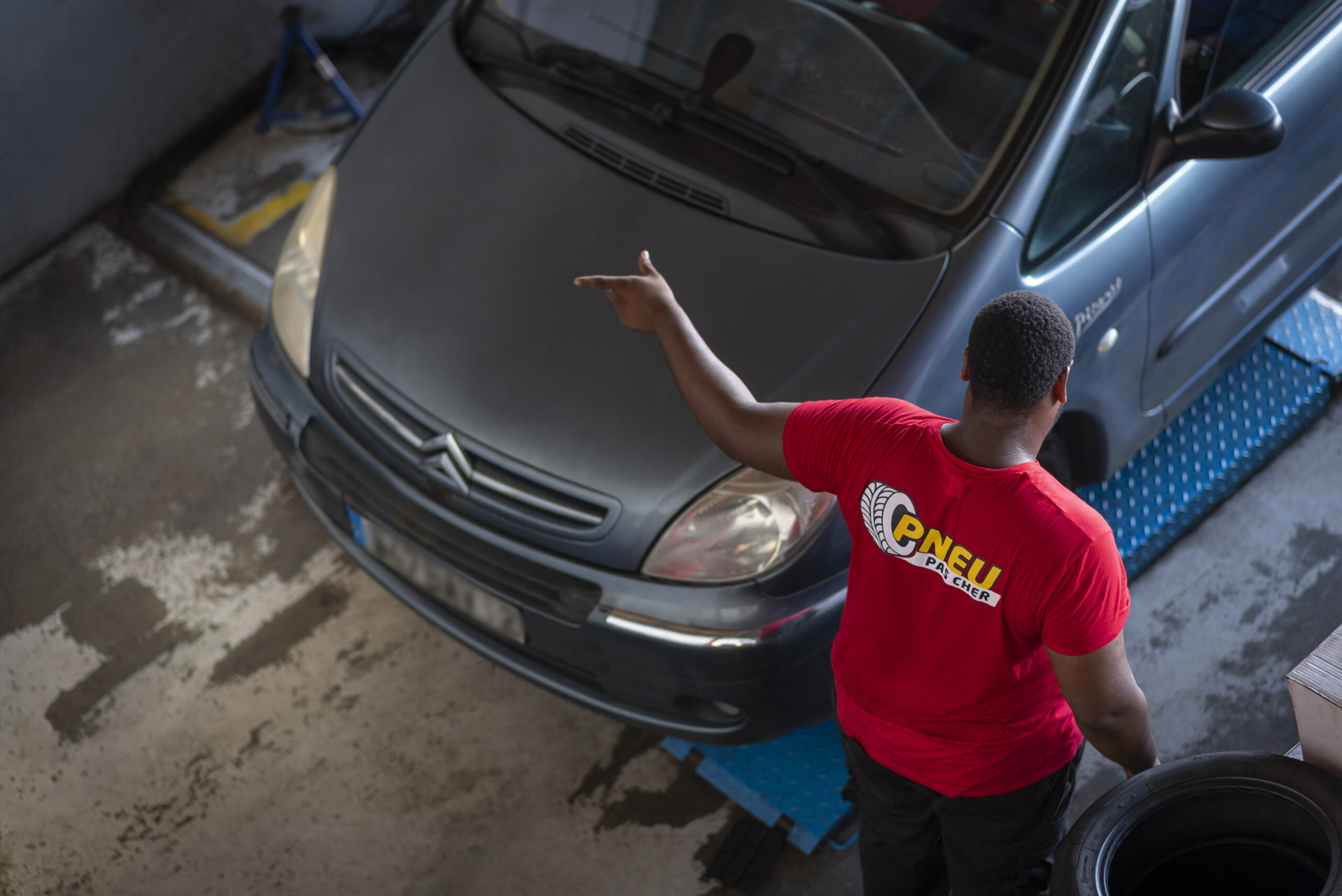 Technicien Pneu Pas Cher en tenue rouge indiquant le capot d'une voiture dans le centre de Place d'Armes pour une intervention batterie